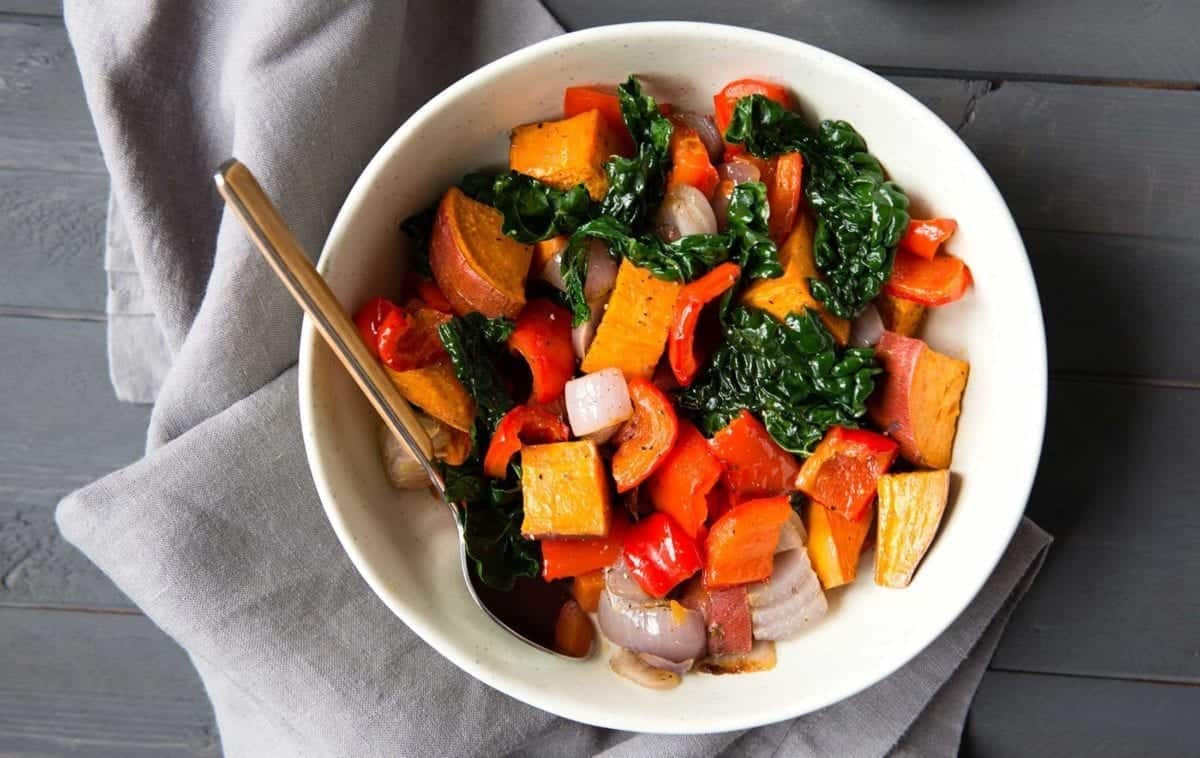 White bowl filled with roasted sweet potatoes, bell peppers, red onions, and kale, served with a spoon on a gray napkin.