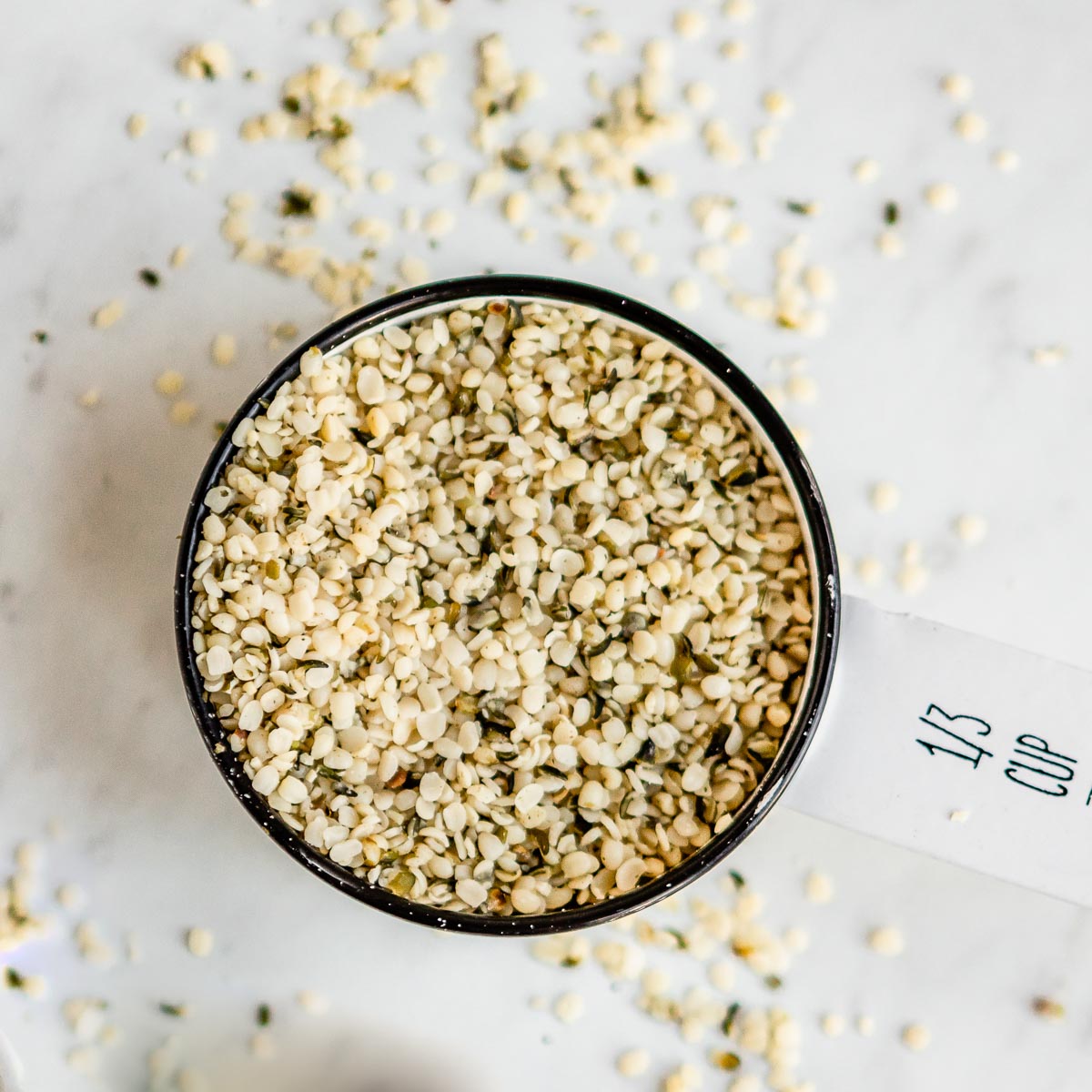 White measuring cup filled with hemp hearts spilling over, ideal for hemp hearts recipes and healthy eating.