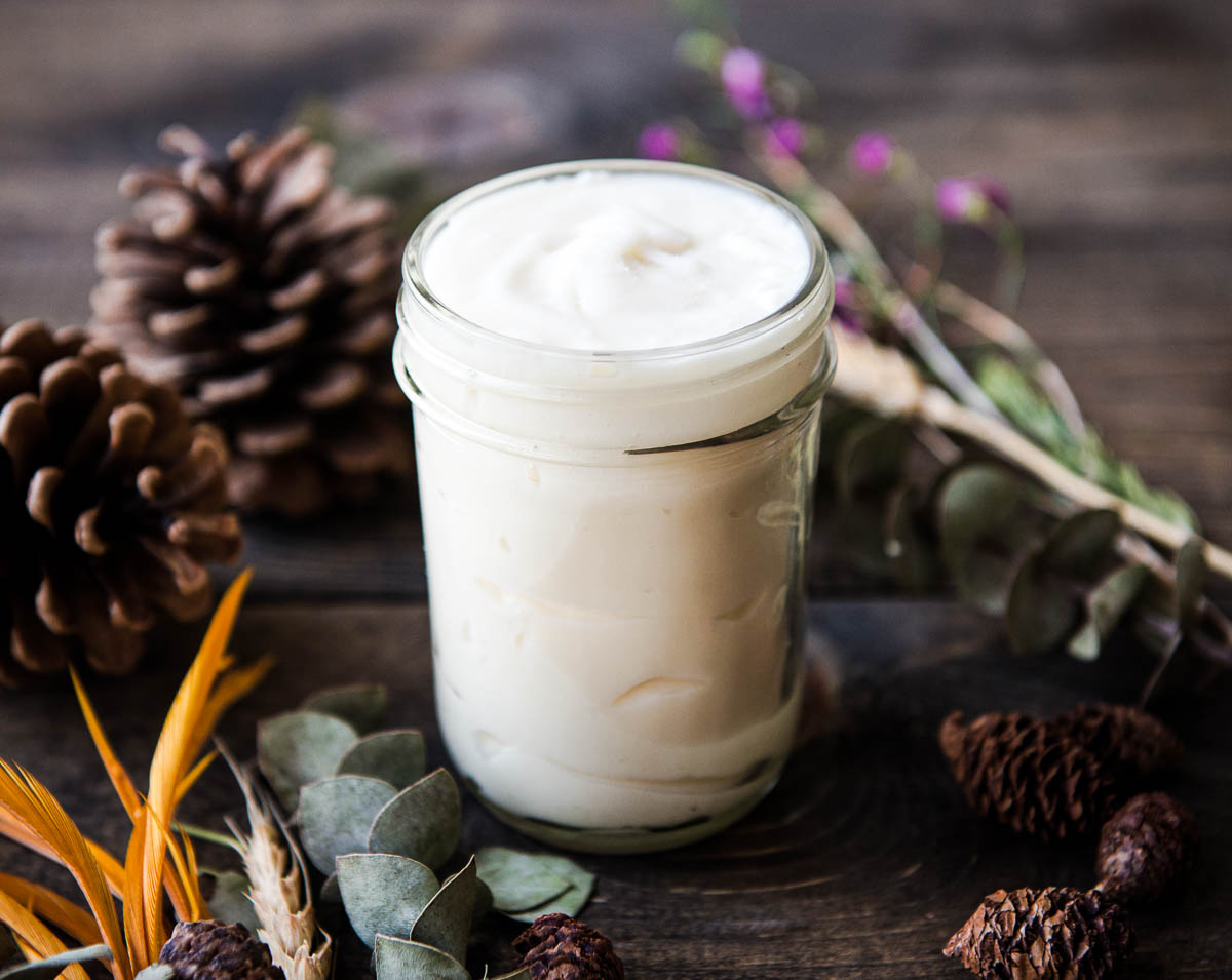 A glass jar of natural homemade deodorant made with coconut oil and baking soda, sitting on a dark woodgrain table, surrounded by pine cones and flowers.