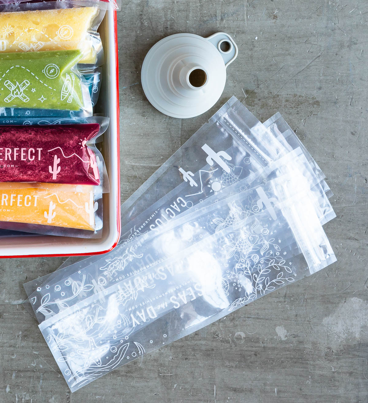 light gray funnel, several popsicle sleeves and a red tray full of colorful popsicles on a gray counter top.