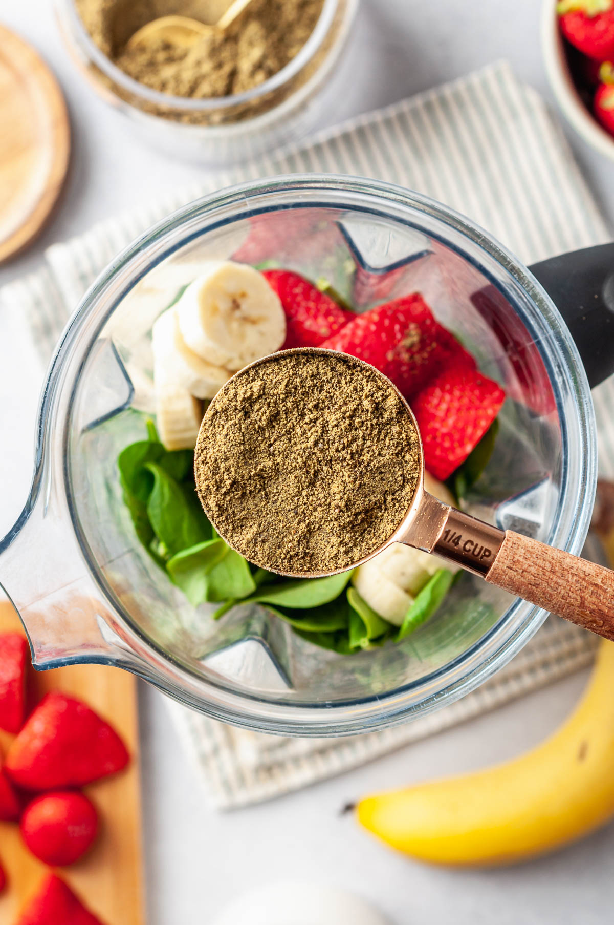 Scoop with best organic protein powder hovering over a blender full of smoothie ingredients: green spinach, banana slices and bright strawberries.