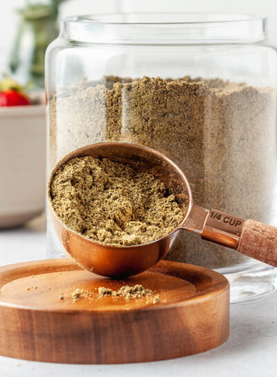 Scoop of homemade protein powder in a 1/4 cup measuring spoon in front of a large glass jar of the full batch of DIY protein powder.