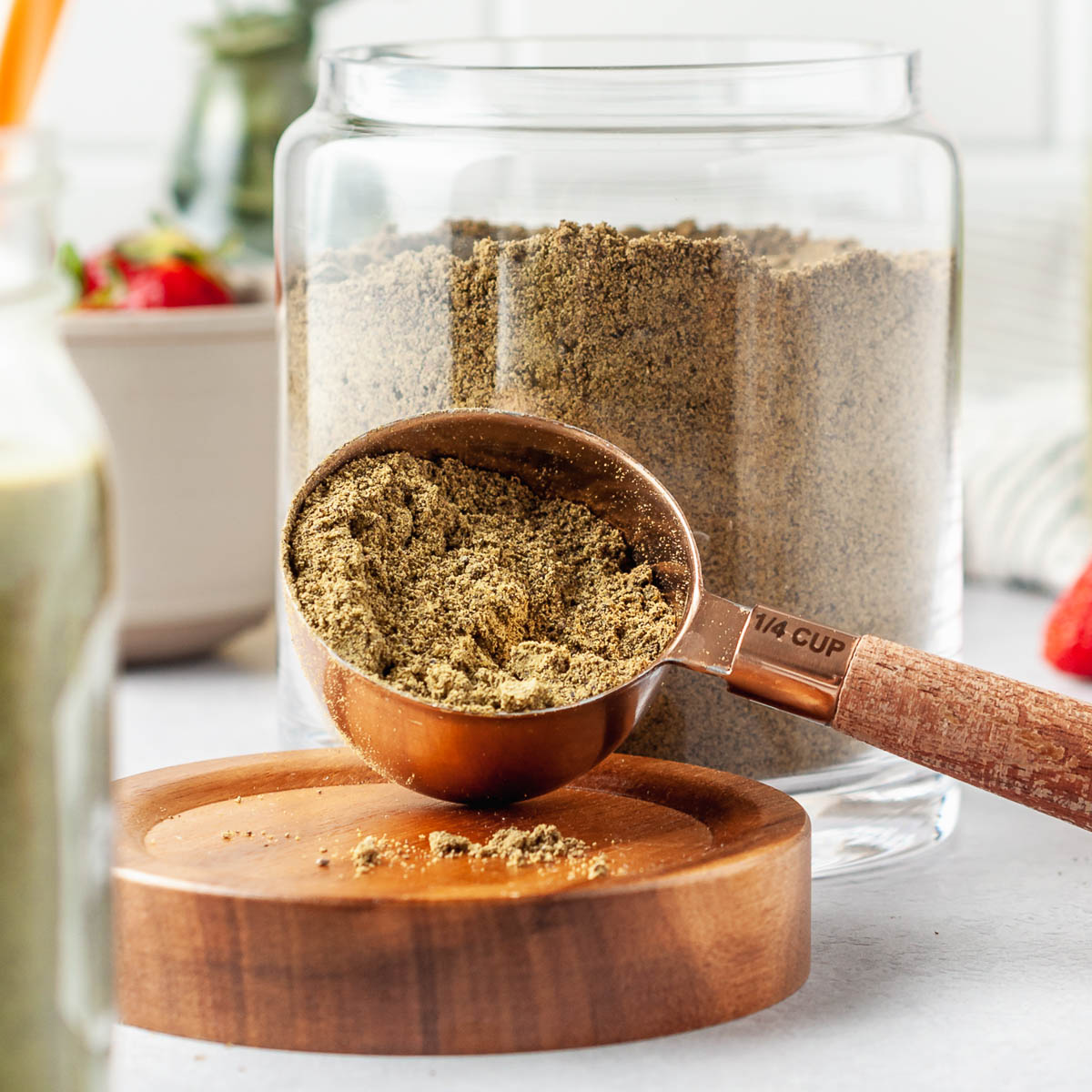 Scoop of homemade protein powder in a 1/4 cup measuring spoon in front of a large glass jar of the full batch of DIY protein powder.