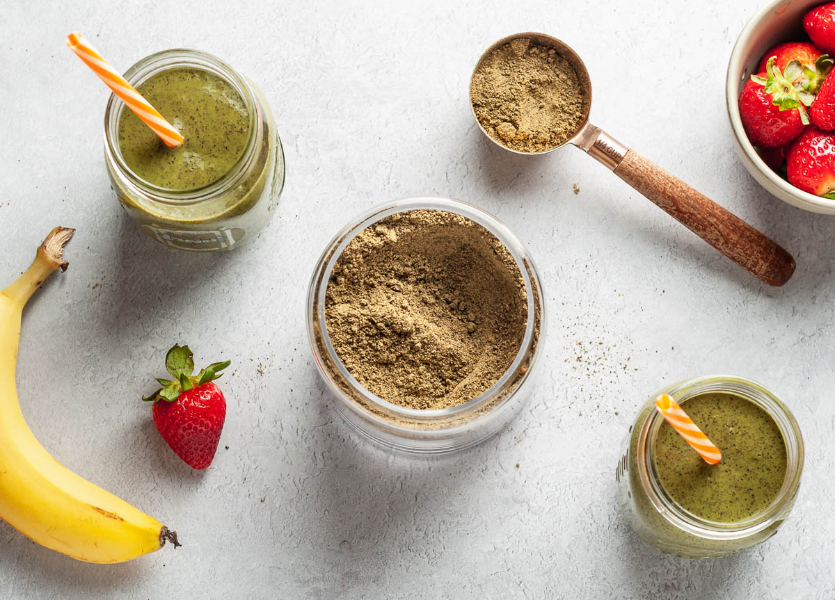 Homemade protein powder in between two glass jars of green smoothies, with orange straws, surrounded by a banana, strawberries, and a scoop of protein powder.