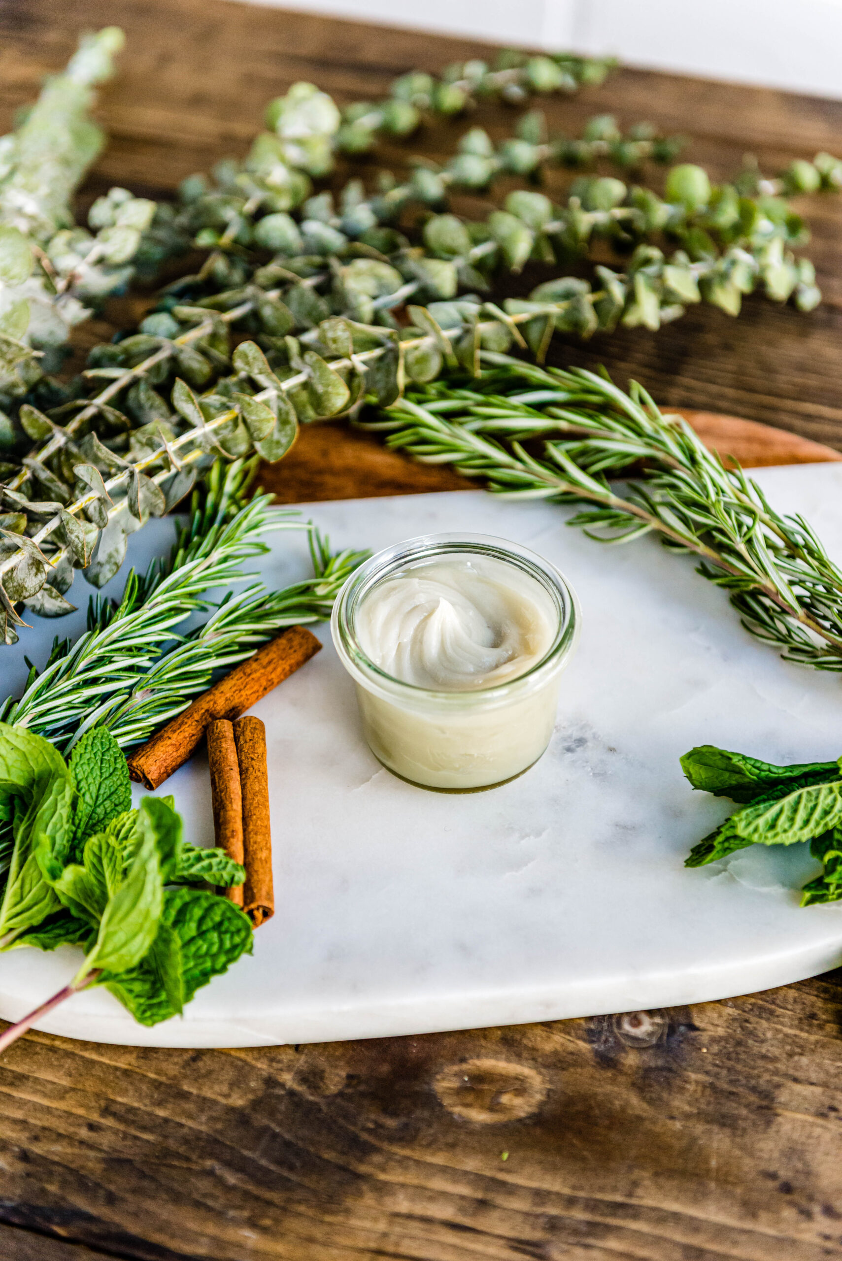 glass jar of natural decongestant on white marble surrounded by fresh herbs and spices.