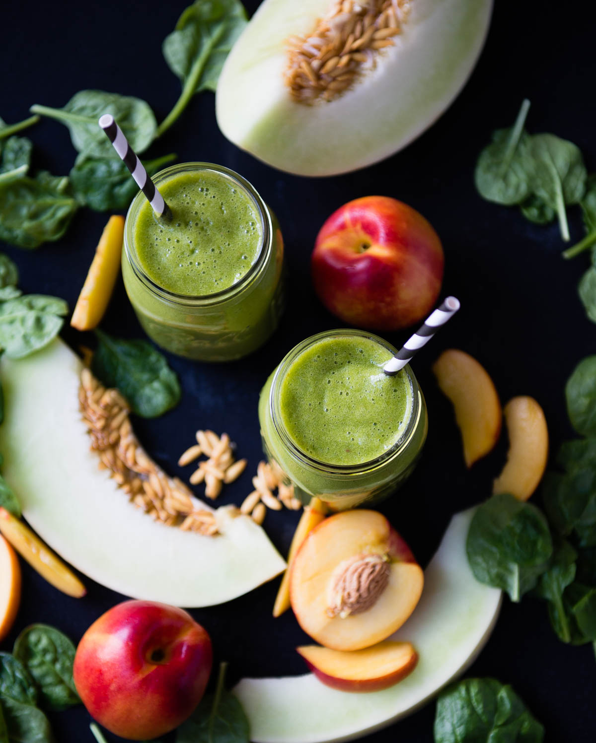 2 green smoothies in glass jars with paper straws on a counter with fresh spinach, honeydew, and nectarines.