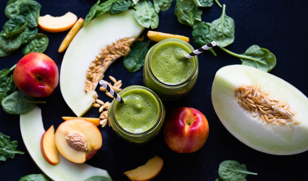 2 green smoothies in glass jars with black and white paper straws on a black counter next to fresh ingredients.