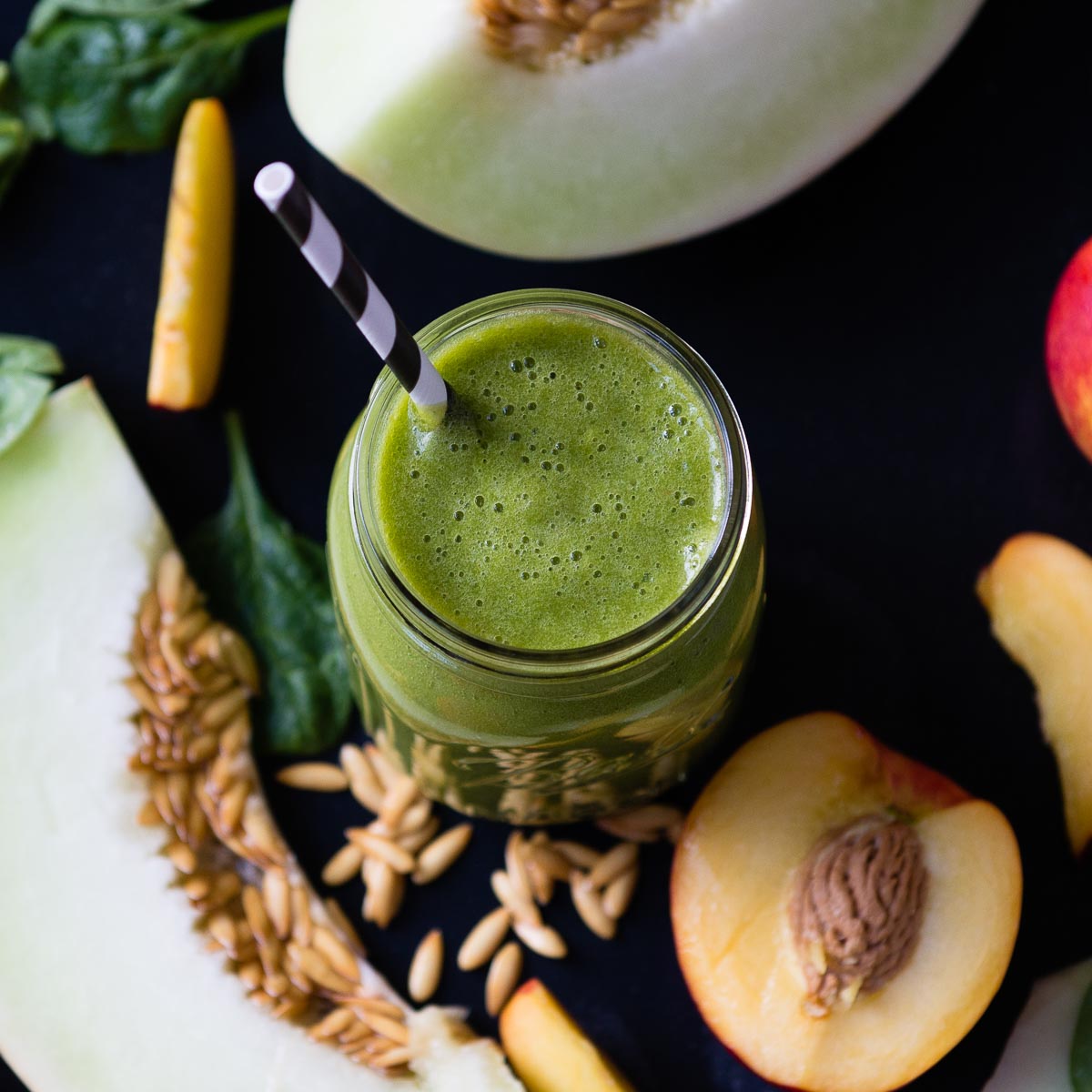 honeydew smoothie in a glass jar with a striped paper straw, next to a nectarine and a sliced honeydew.