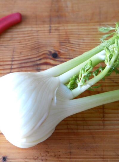 fresh fennel on a wooden cutting board next to a paring knife with a red handle.