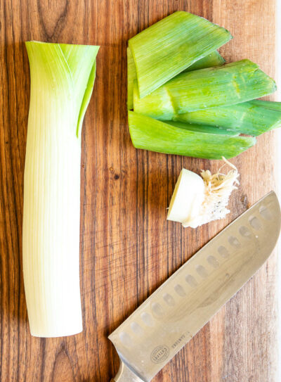 partially cut leek on a wooden cutting board next to a chef's knife.