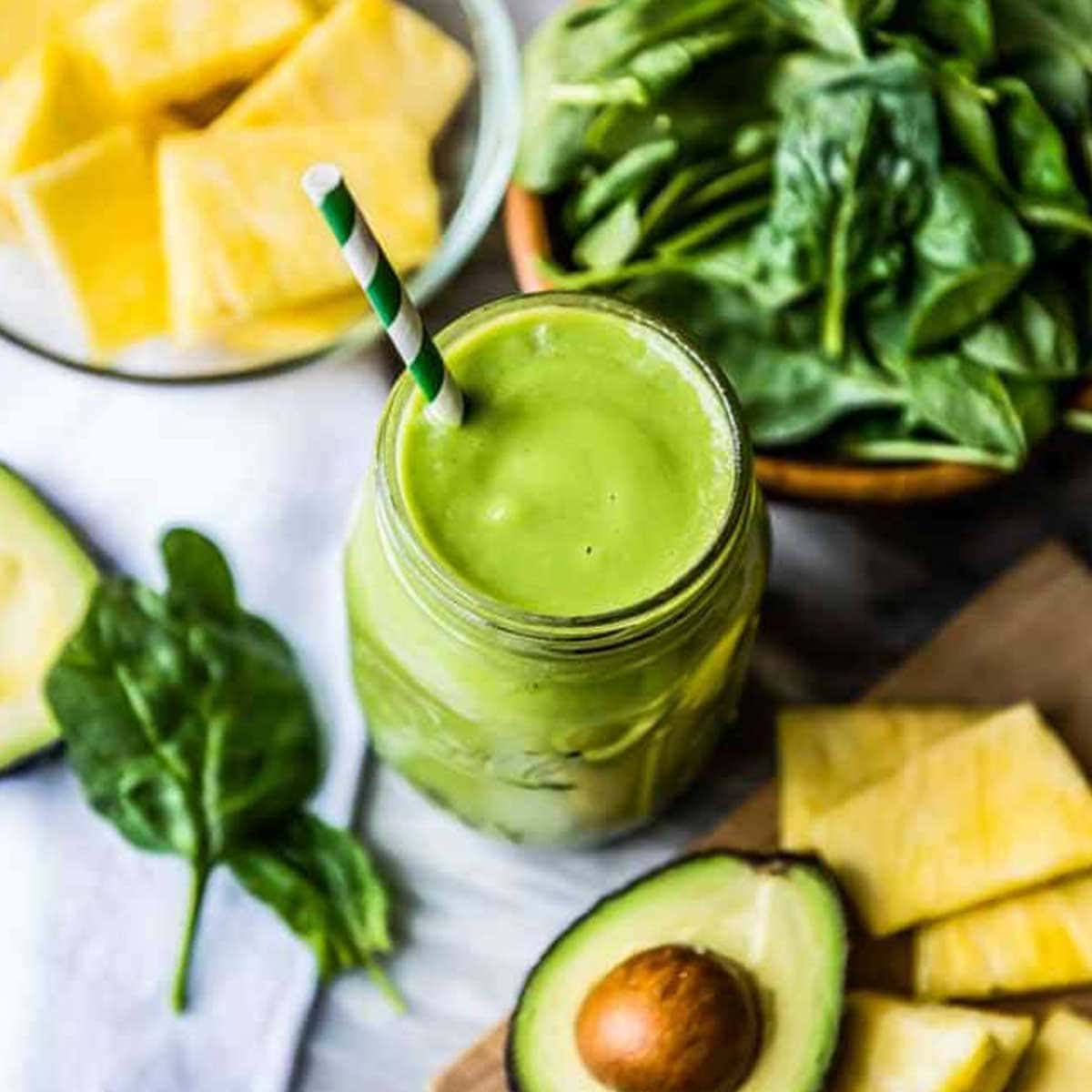 Green clear skin smoothie in a mason jar with a paper straw, on a counter with avocado, pineapple and spinach.