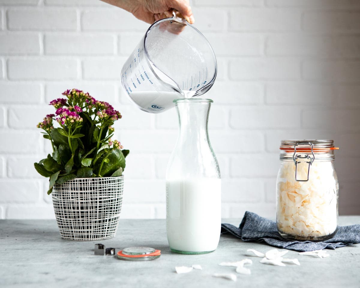 pouring homemade coconut milk into a glass pitcher on a counter with coconut flakes in a jar.