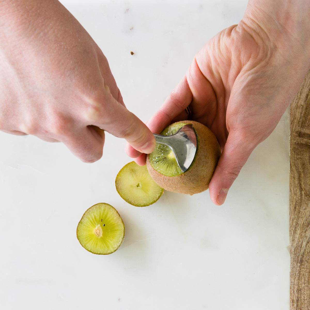 2 hands using a spoon to slip between the skin and flesh of a kiwi.