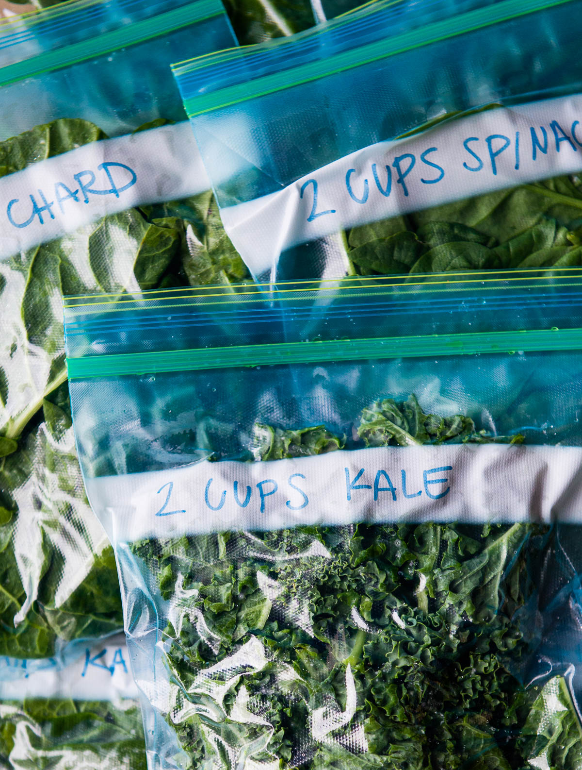 several ziploc bags containing different kinds of leafy greens, ready to be frozen. The one on top is labeled 2 cups kale.