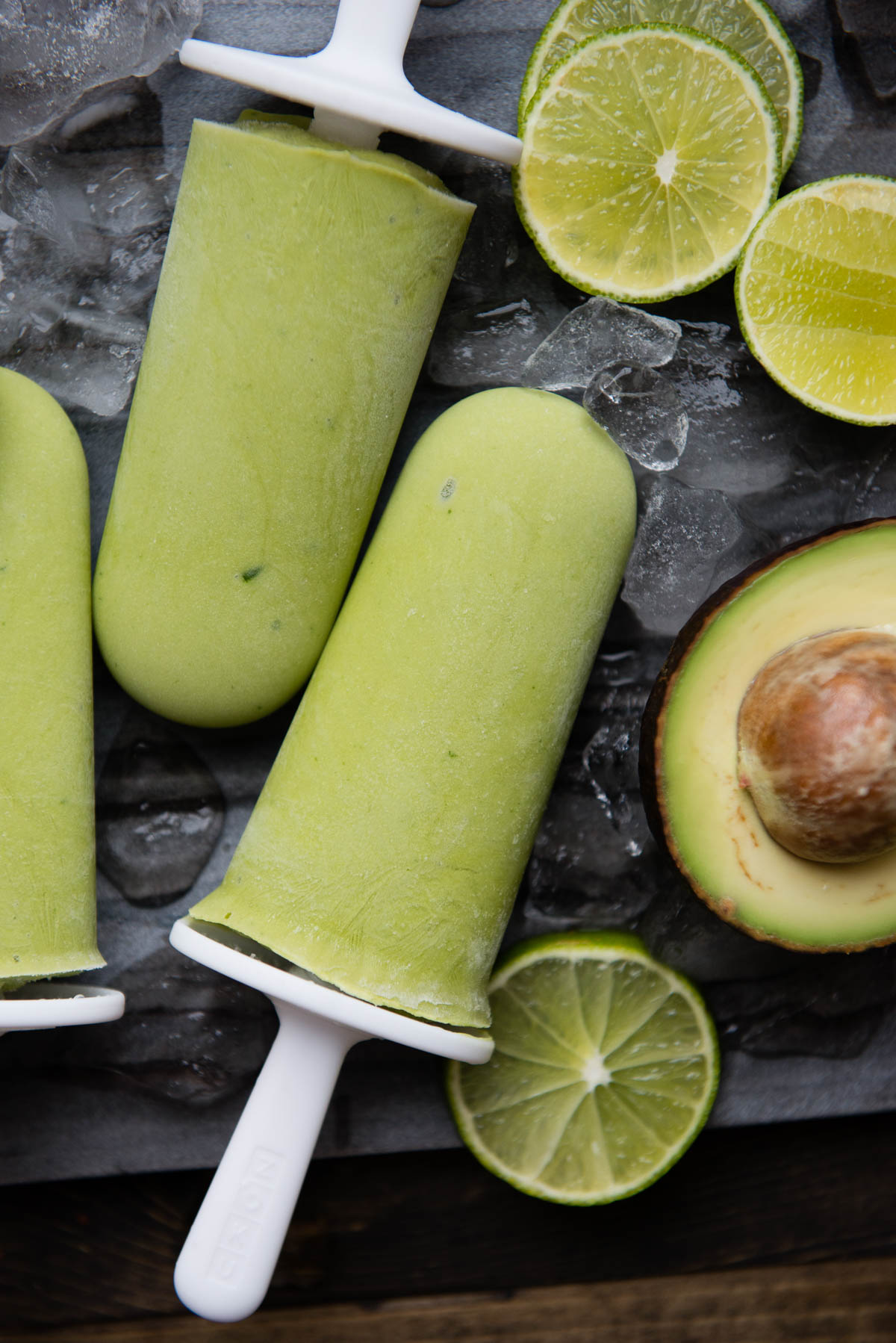 green popsicles on white sticks on a stone slab with ice cubes and lime slices and avocado.