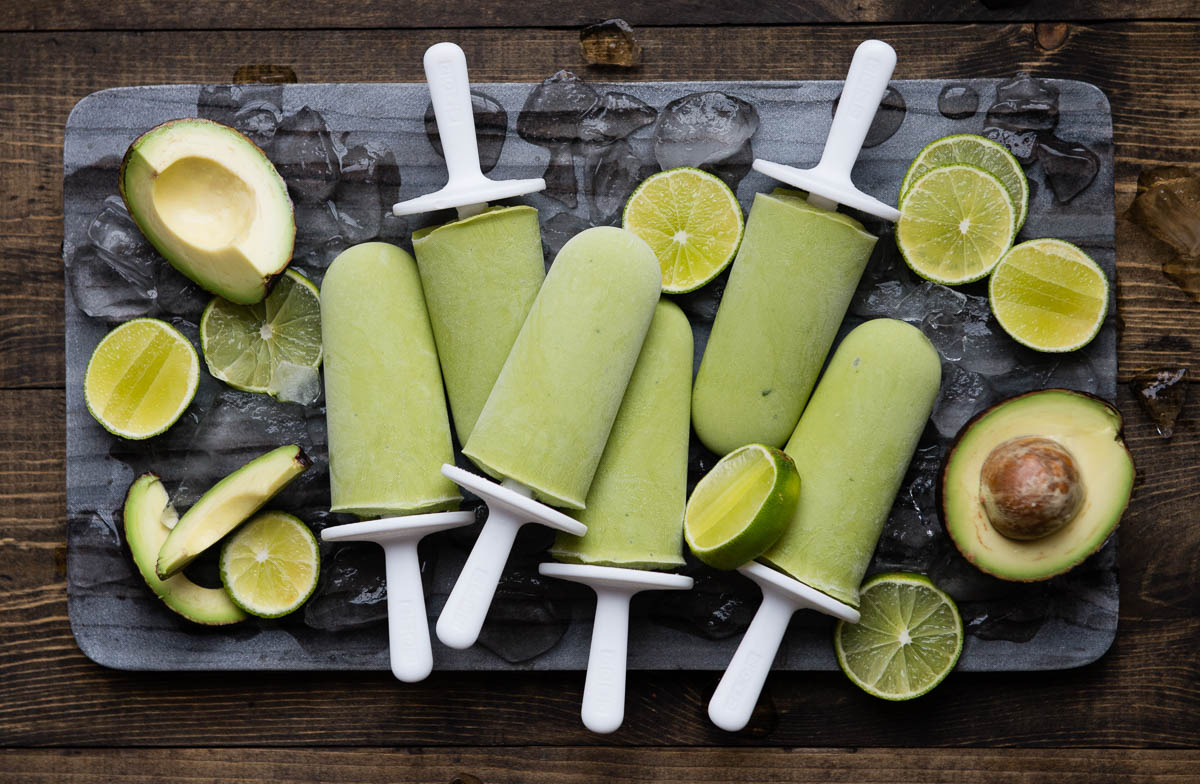 6 lime popsicles on white sticks on a stone slab with ice cubes and lime slices.