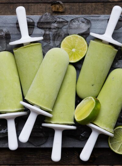 6 lime popsicles on white sticks on a stone slab with ice cubes and lime slices.