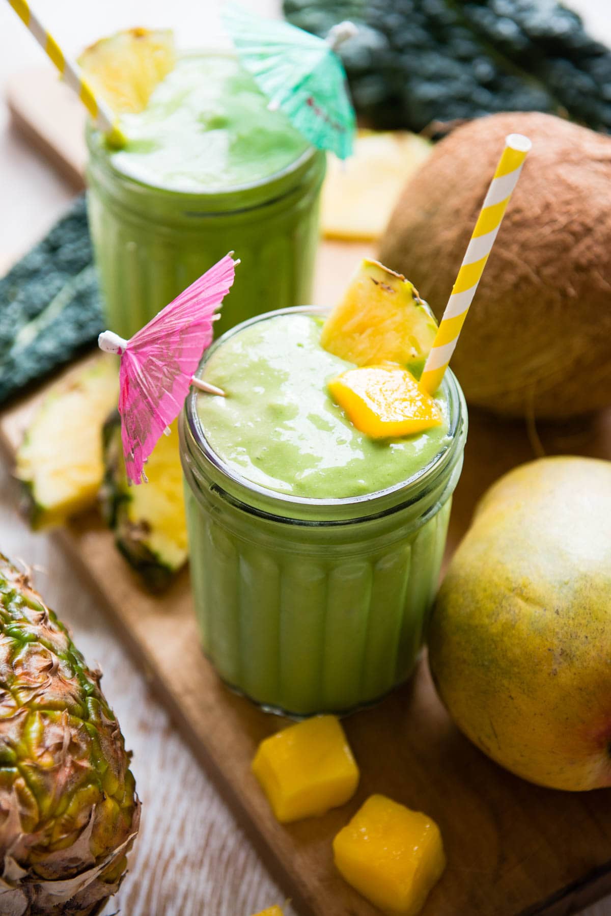 2 jars of kale mango smoothies in glasses with colorful straws, umbrellas, and whole fruits around them.