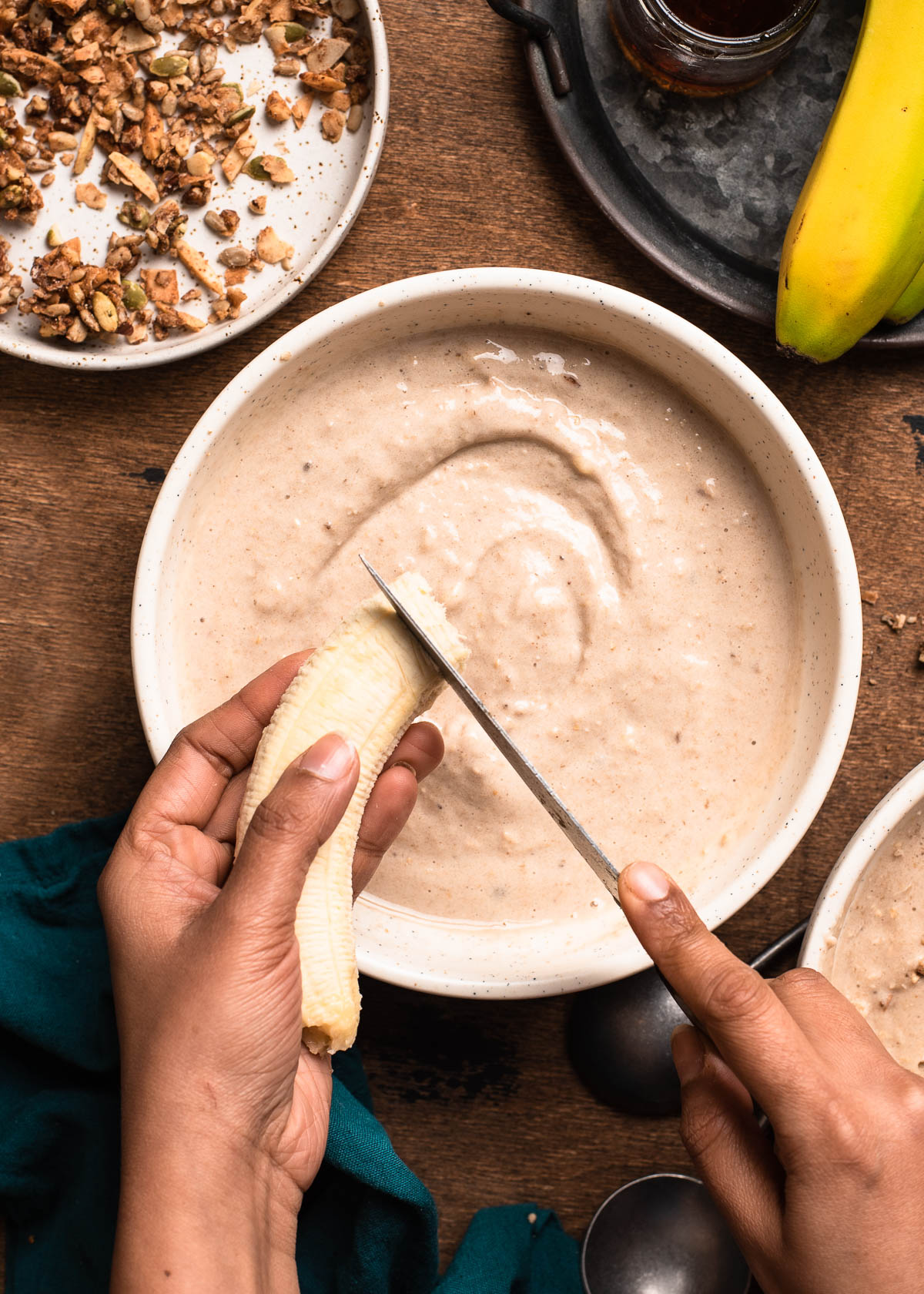 2 hands slicing a banana over a smoothie bowl.