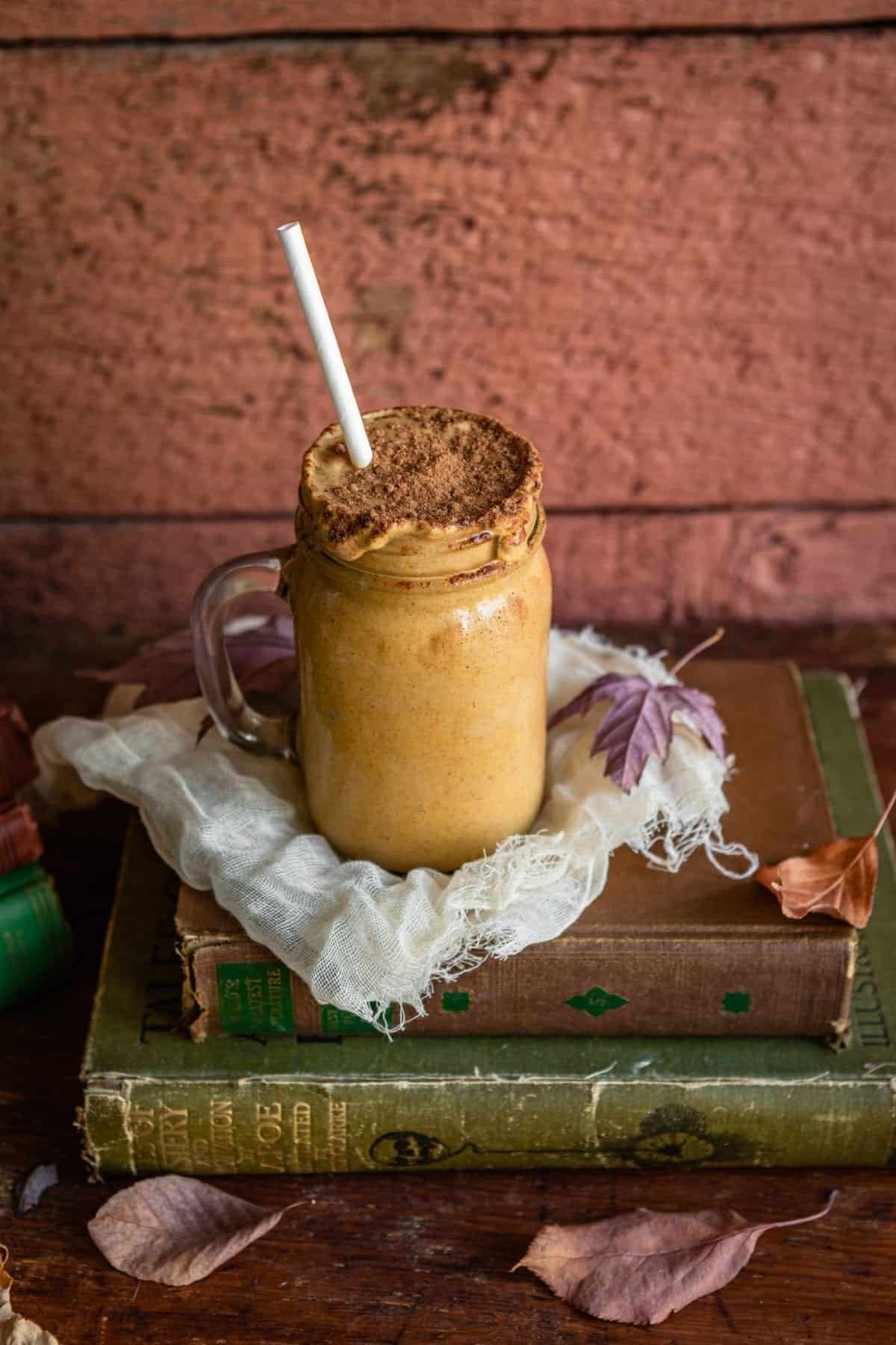 maple pumpkin fall smoothie in a glass mug with a paper straw on top of cheese cloth and a stack of antique books.