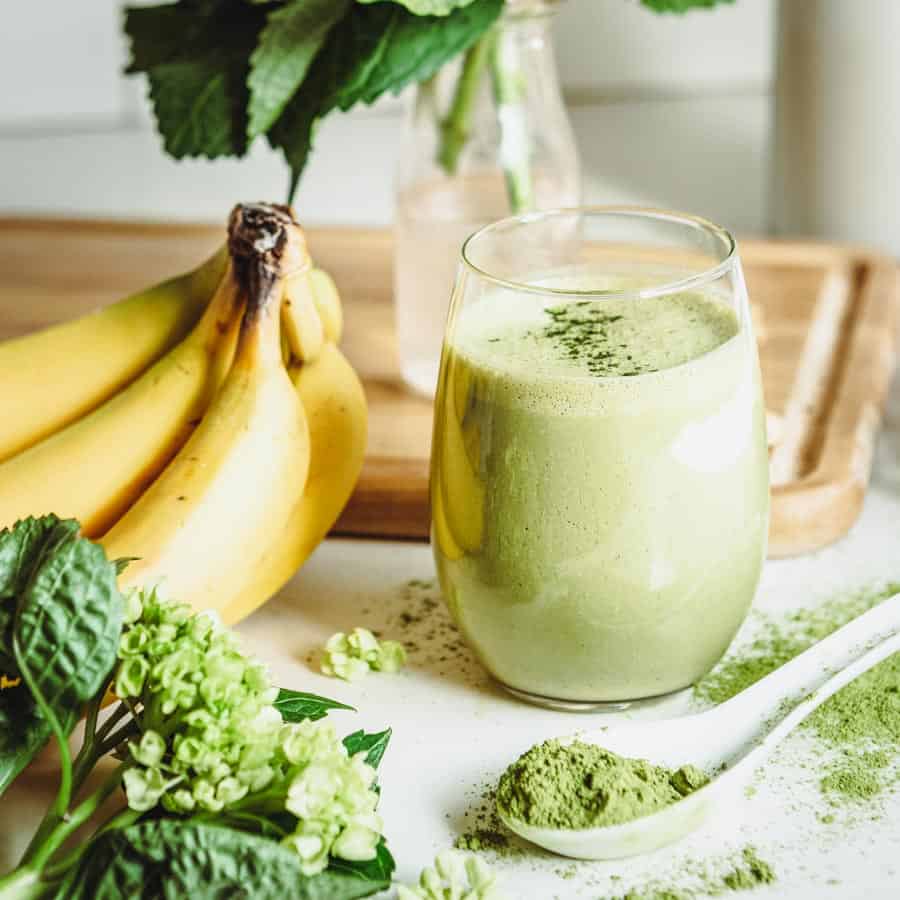 matcha smoothie in a round glass on a white counter top surrounded by fresh ingredients.