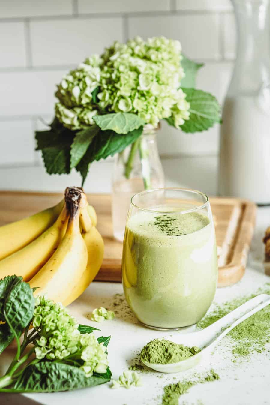 matcha smoothie in a round glass surrounded by bananas, matcha powder and a hydrangea.