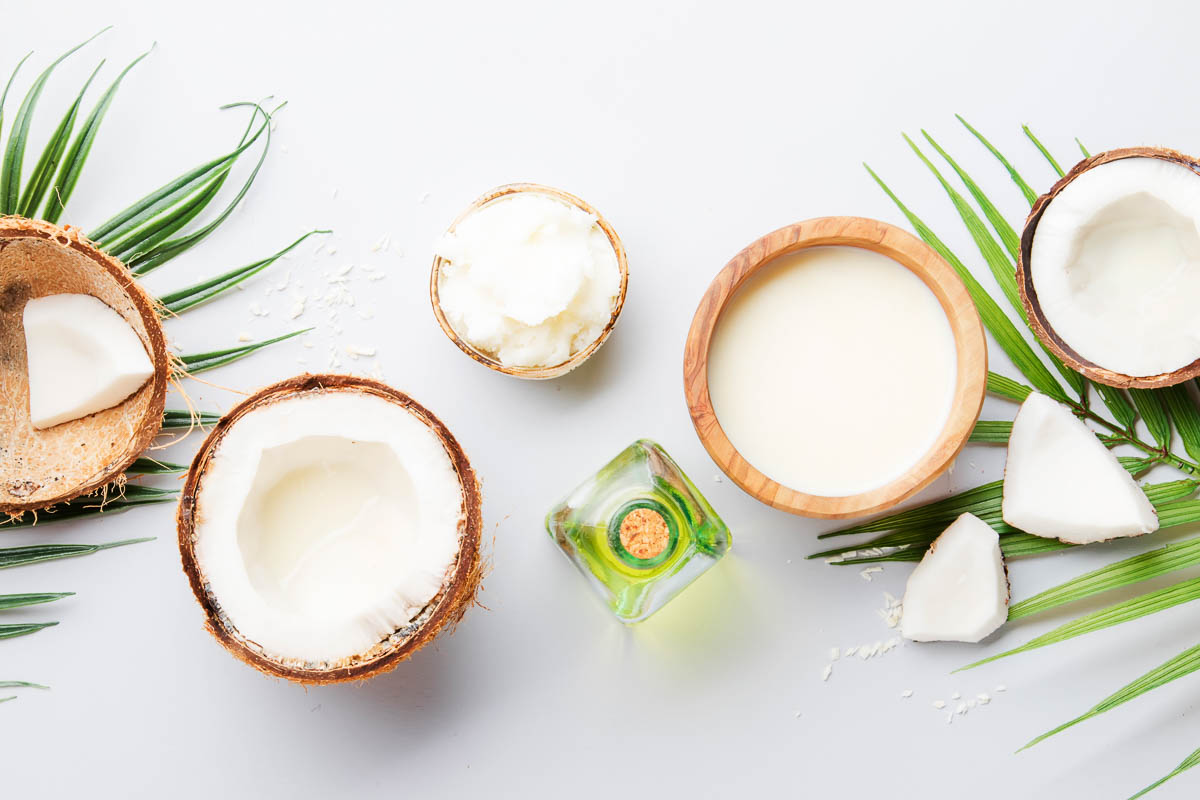 white counter with sliced white coconuts next to a bowl of solid coconut oil and a glass bottle of liquid coconut oil.