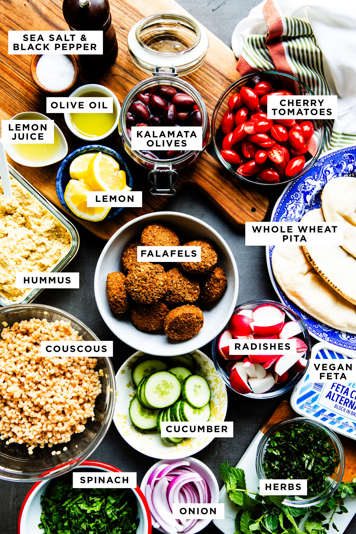 labeled ingredients for a Mediterranean Falafel bowl including sea salt and black pepper, olive oil, cherry tomatoes, Kalamata olives, lemon juice, lemon, hummus, falafel, whole wheat pita, couscous, radishes, cucumber, spinach, onion and herbs.