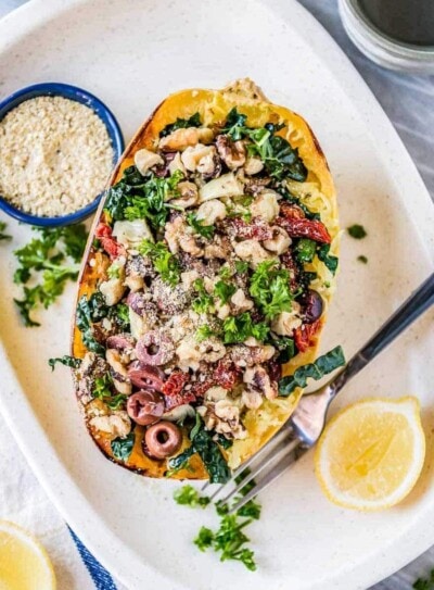 oven roasted spaghetti squash topped with olives, vegan parmesan, kale and artichokes on a white platter with sliced lemons.