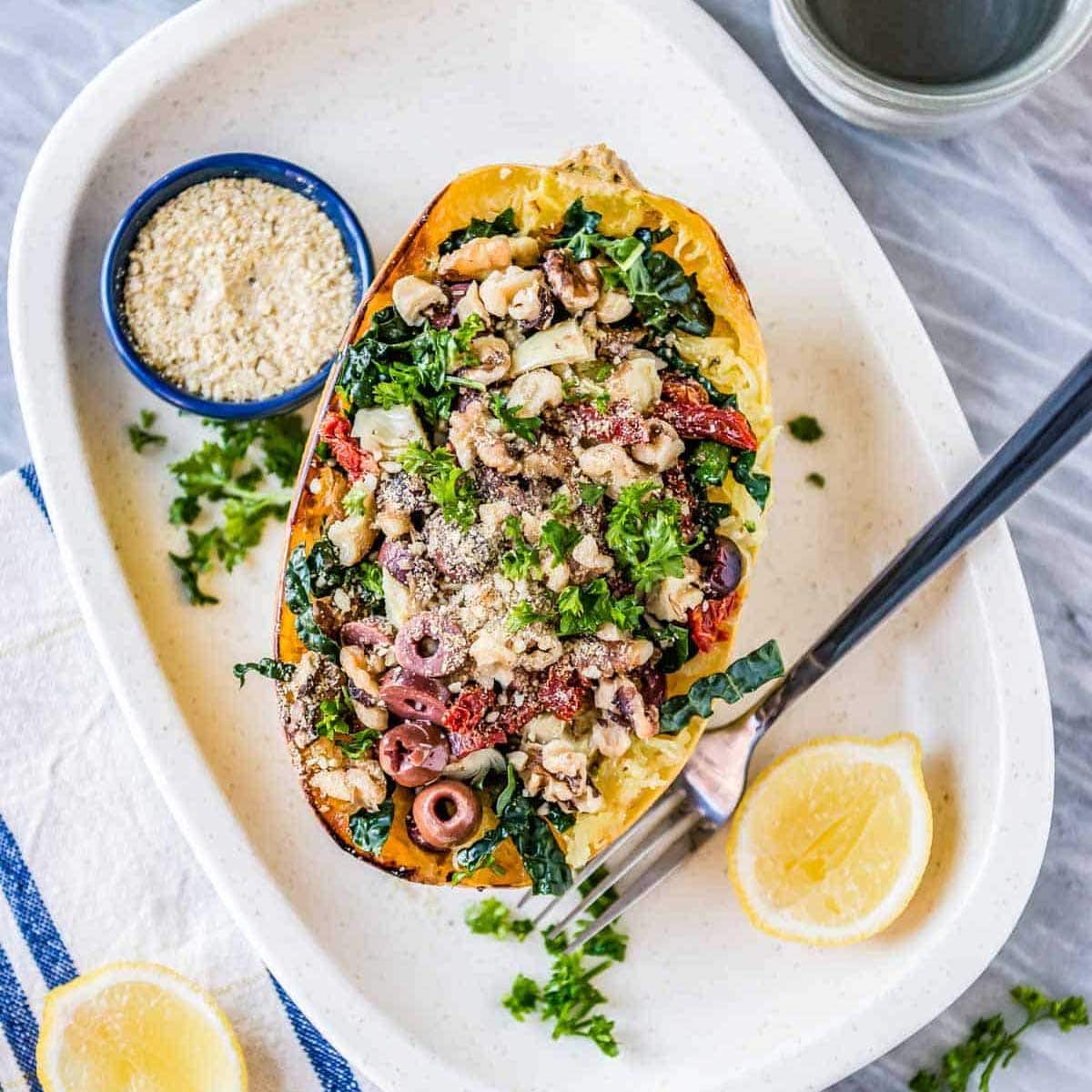 oven roasted spaghetti squash topped with olives, vegan parmesan, kale and artichokes on a white platter with sliced lemons.