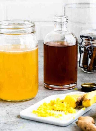 yellow metabolism tea in clear glass jar next to a glass jar of apple cider vinegar on a counter with fresh sliced ginger and turmeric.