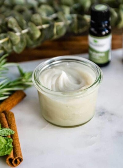 Natural homemade vapor rub in glass jar surrounded by essential oils and herbs.