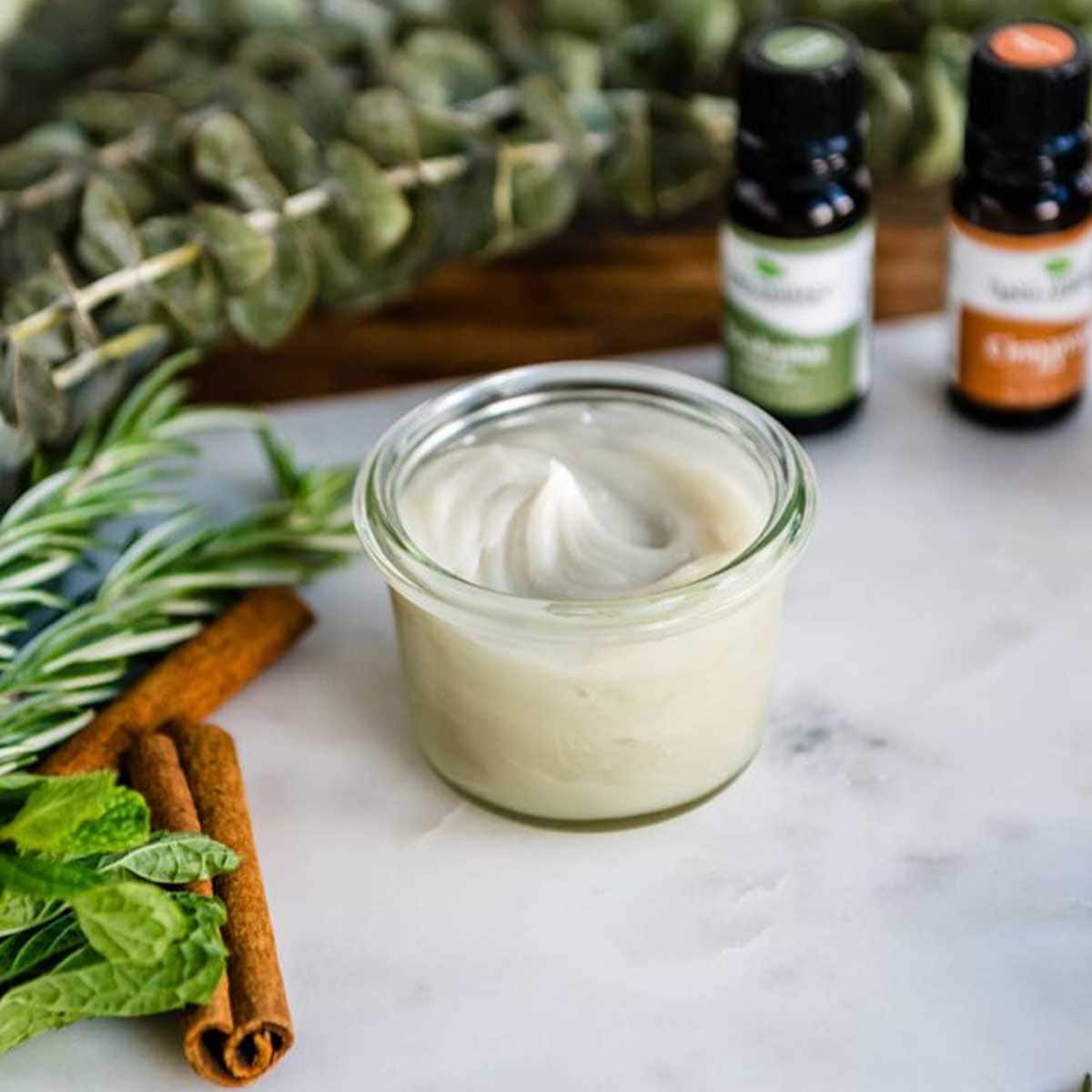 Natural homemade vapor rub in glass jar surrounded by essential oils and herbs.