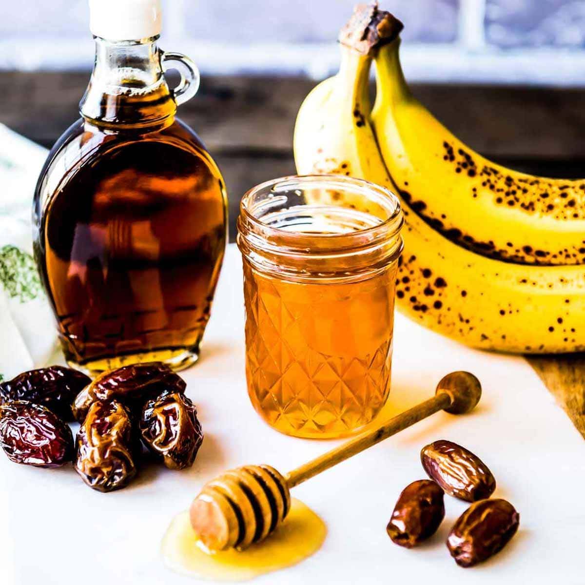 examples of natural sweeteners including bananas, honey in a glass jar, maple syrup and dates.