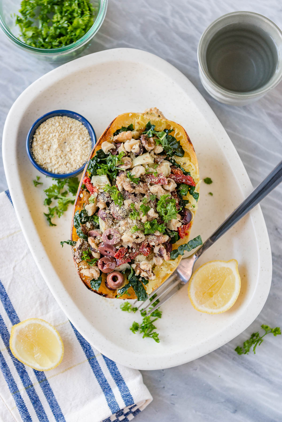 oven roasted spaghetti squash topped with olives, vegan parmesan, kale and artichokes on a white platter with sliced lemons.