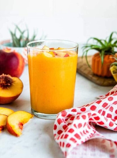 glass of peach smoothie topped with fresh peaches, on a white countertop with whole and sliced peaches.