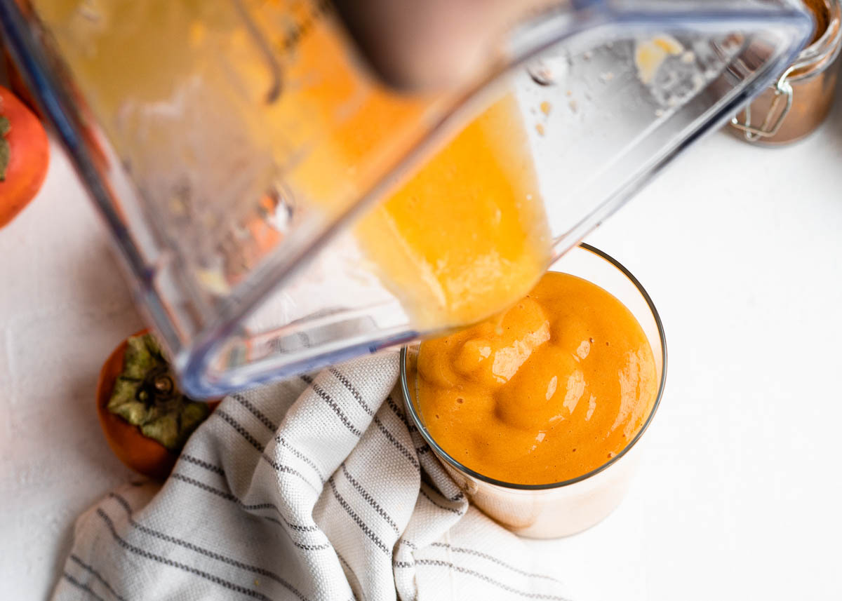 pouring an orange fruit smoothie from a blender container into a glass sitting on a white countertop.