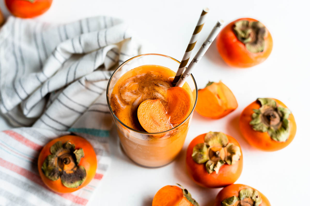 glass of yogurt persimmon smoothie topped with fresh persimmons and 2 paper straws, on a white counter surrounded by persimmons.