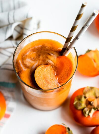 glass of yogurt persimmon smoothie topped with fresh persimmons and 2 paper straws, on a white counter surrounded by persimmons.