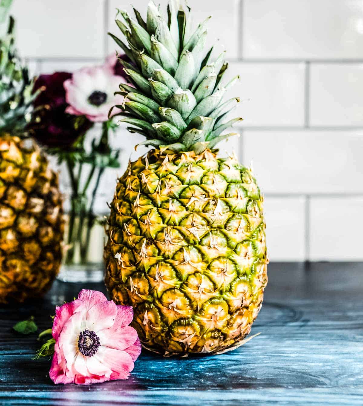 A whole pineapple next to a pink and white flower on a blue countertop, perfectly ready to make the best tropical smoothie.