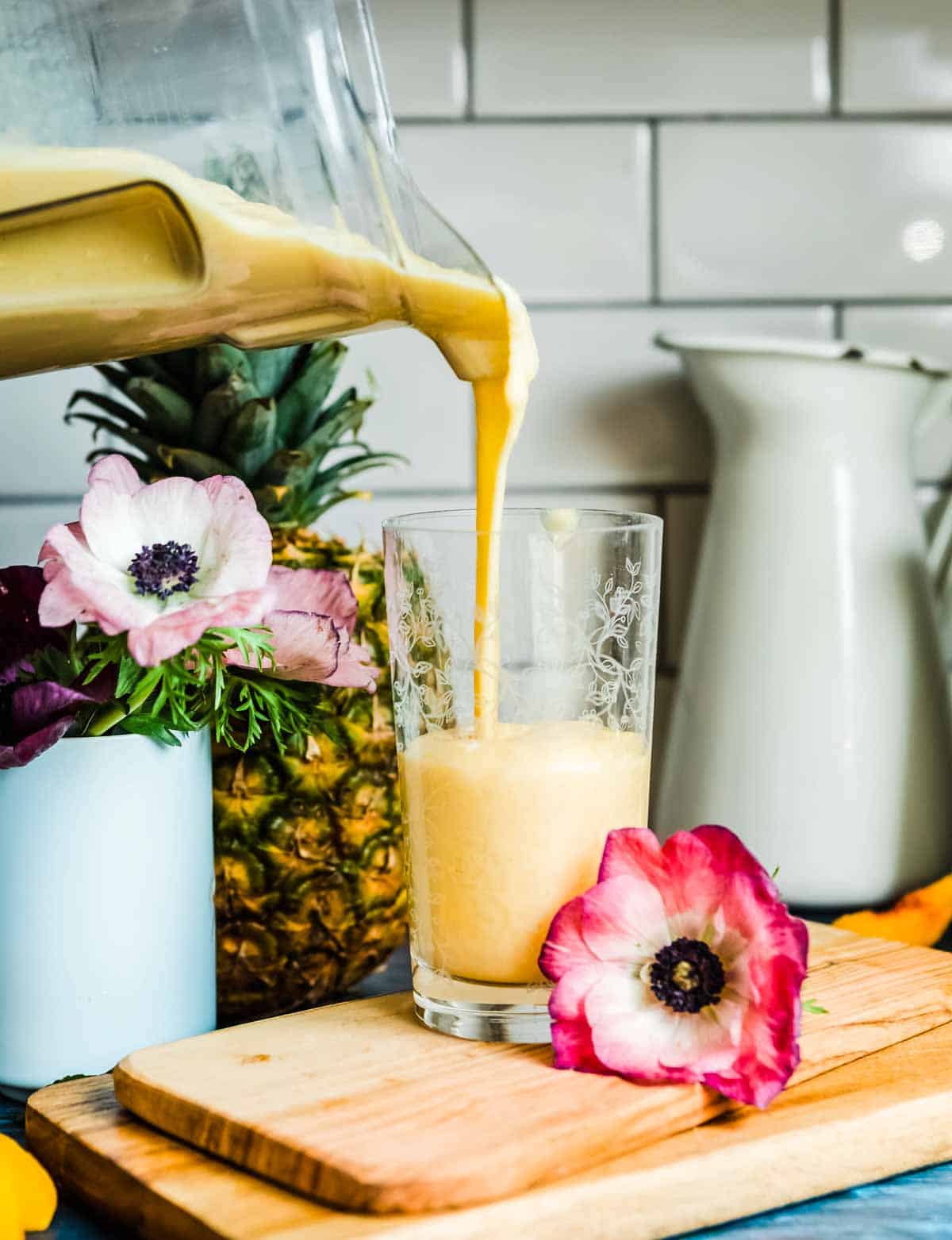 Pouring bright yellow pineapple smoothie liquid into a glass from a blender container.