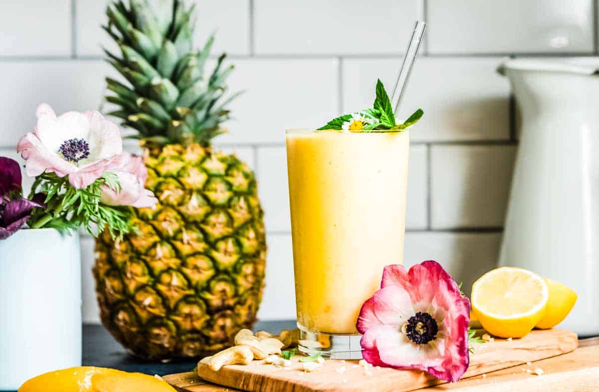 Bright yellow pineapple smoothie in a glass with a glass straw on a counter top, along with ingredients and flowers.