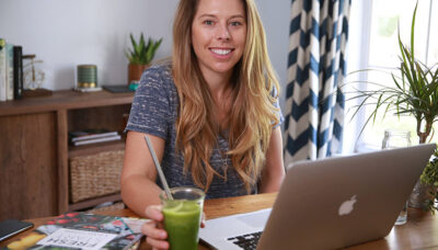 Jen at her desk and computer with a smile and green smoothie in hand.