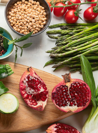 Fresh ingredients including tomatoes, asparagus, pomegranate, cucumber slices, and bok choy spread out on white counter top.