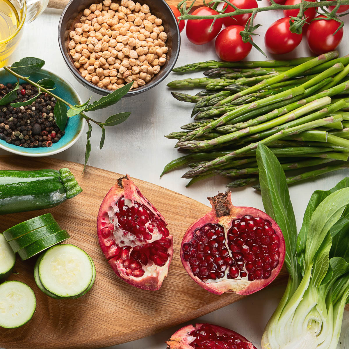 Fresh ingredients including tomatoes, asparagus, pomegranate, cucumber slices, and bok choy spread out on white counter top.