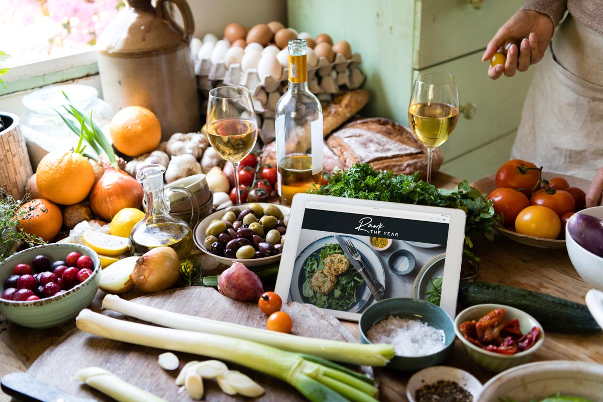 wooden counter covered with fresh tomatoes, olives, berries, sea salt, leeks, onions, oranges, eggs, 2 wine glasses with white wine and a tablet in the center that has a Rawk the Year page pulled up.