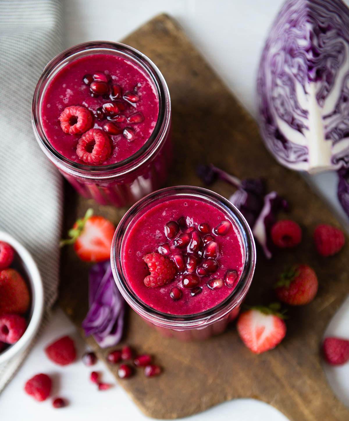 two glass jars full of pomegranate smoothies topped with pomegranate arils and raspberries, sitting on a wooden cutting board.