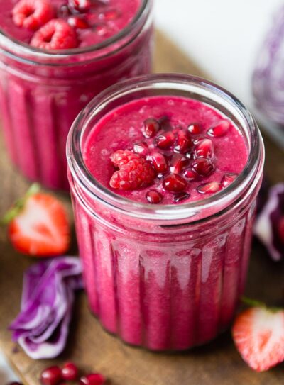 two glass jars full of pomegranate smoothies topped with pomegranate arils and raspberries, sitting on a wooden cutting board.