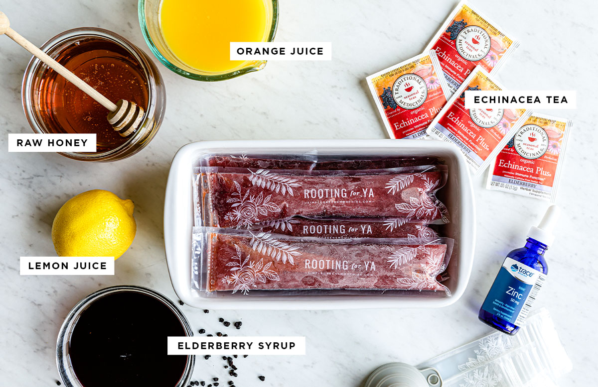 Ingredients for popsicles for sore throats laid out on a white counter with labels: orange juice, echinacea tea, elderberry syrup, lemon juice and raw honey.