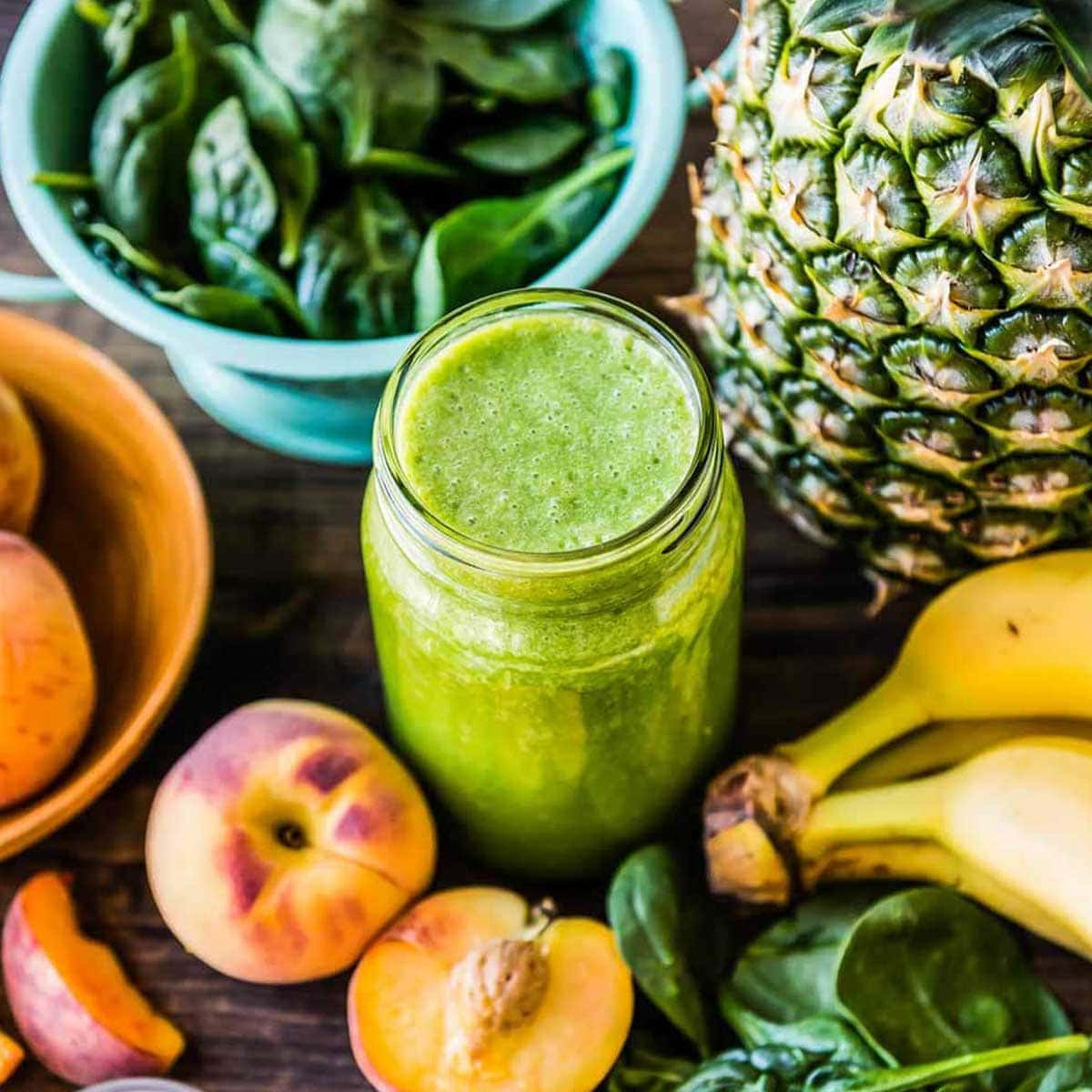 Green pre-workout smoothie in a glass jar surrounded by fresh spinach, bananas, peaches, and pineapple, perfect for a pre-workout smoothie recipe.